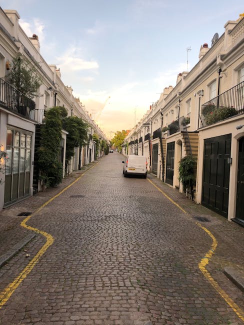 A narrow cobblestone street lined with white terraced houses in a residential area, with small front gardens and balconies, and greenery such as potted plants and trees. A white van is parked along the curb in the middle distance, and the street is flanked by black and white painted borders designating parking or loading zones. The sky above is partly cloudy with a hint of sunset light, casting a soft illumination over the scene. The environment appears quiet and typical of an affluent urban neighbourhood, with no visible rubbish or debris, suggesting that private waste collection services like those provided by Rubbish Removal Westminster may be used for on-site clearance or alternative waste handling within these properties.