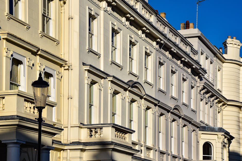 This image depicts a section of a historic multi-storey building with a cream-colored facade, detailed with decorative moldings and window surrounds. The structure features multiple large, rectangular sash windows with white frames, some of which have decorative pediments above them. A black vintage-style street lamp is positioned on the sidewalk in the foreground, standing upright with a clear glass enclosure for the bulb. The building's exterior walls have a textured surface, and the cornices and window sills are accentuated with dark material, contrasting with the light-colored facade. To the right, part of a rounded turret-like extension is visible, with a chimney stack on the rooftop. The sky is clear and blue, providing natural sunlight that highlights the architectural details. This scene suggests an urban environment, potentially near residential or commercial premises, where such detailed facades may be repaired or maintained, or where private property owners might arrange for local or independent waste clearance services to manage building renovation or maintenance debris. Rubbish Removal Westminster offers services relevant to such contexts, ensuring proper waste handling for older buildings during clearance projects or property maintenance tasks.