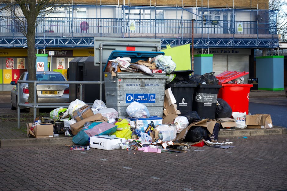 A large collection of discarded waste and rubbish bags accumulated around two wheelie bins on a paved area in an urban setting. The waste includes cardboard boxes, plastic bags, paper packaging, and miscellaneous paper and plastic debris, some of which spill onto the brick pavement in front of the bins. The bins are situated beside a waist-high metal railing, with one black and one black and red bin, indicating mixed waste and recycling separation. Behind the waste, there is a parked silver car with a visible registration plate. In the background, a building under renovation is covered with blue scaffolding and safety netting, with storefronts on the ground floor featuring various signage. The scene is illuminated with natural daylight, suggesting daytime, and the environment appears to be in a public parking or loading area, highlighting the importance of private waste collection services like those offered by Rubbish Removal Westminster for alternative rubbish disposal options.