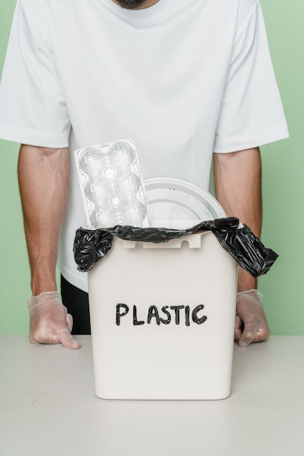 A person wearing a white t-shirt and transparent plastic gloves stands behind a white plastic rubbish bin labeled 'PLASTIC.' The bin's lid is slightly open, with a black trash bag draped over the edge, partially inside the container. Resting on top of the bin is a clear plastic tray with multiple circular depressions, typical of food or ice packaging. The background is a plain, light green wall, providing a neutral setting for the scene. This image highlights waste segregation practices, with a focus on recyclable plastics, corresponding to alternative rubbish disposal methods such as private collection or on-site clearance services often handled by waste management specialists like Rubbish Removal Westminster, operating in London boroughs including Westminster.