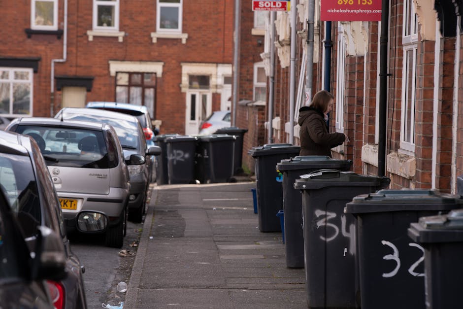 A large collection of discarded waste and rubbish bags accumulated around two wheelie bins on a paved area in an urban setting. The waste includes cardboard boxes, plastic bags, paper packaging, and miscellaneous paper and plastic debris, some of which spill onto the brick pavement in front of the bins. The bins are situated beside a waist-high metal railing, with one black and one black and red bin, indicating mixed waste and recycling separation. Behind the waste, there is a parked silver car with a visible registration plate. In the background, a building under renovation is covered with blue scaffolding and safety netting, with storefronts on the ground floor featuring various signage. The scene is illuminated with natural daylight, suggesting daytime, and the environment appears to be in a public parking or loading area, highlighting the importance of private waste collection services like those offered by Rubbish Removal Westminster for alternative rubbish disposal options.