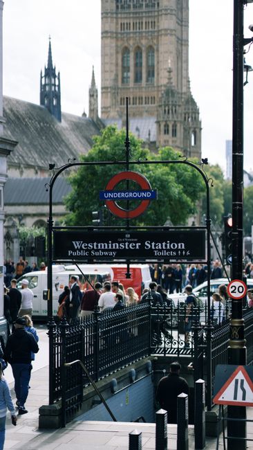The image depicts the entrance to Westminster Underground Station in central London, with a prominent black metal sign featuring the blue, red, and white roundel of the London Underground above the station name 'Westminster Station' and directions for public toilets. Surrounding the entrance are groups of pedestrians, some walking and others waiting or standing near the black metal fencing that lines the station stairs. Behind the station entrance, there is a busy street with vehicles, including cars and a red double-decker bus, in motion. In the background, the historic architecture of Westminster Abbey, with its tall spire and detailed stonework, rises against a cloudy sky, contrasting with the modern urban environment in the foreground. The scene captures a typical bustling day outside Westminster Station, highlighting the area's mix of transportation, historic landmarks, and public activity, relevant to discussions of private or alternative waste handling services near popular transport hubs managed by companies like Rubbish Removal Westminster.
