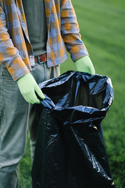 A person wearing a plaid shirt with yellow, blue, and gray tones, as well as green rubber gloves, is holding open a large black rubbish bag made of shiny plastic material. The individual is standing outdoors on a grassy area with a blurred green background, indicating a park or garden setting. The person’s torso and hands are visible, with the bag positioned in front of their waist. The black rubbish bag is partially open, empty, and ready for waste disposal, suggesting an activity related to waste collection or rubbish removal. The scene reflects environmentally conscious waste handling practices, possibly as part of a private rubbish clearance service, with a focus on environmentally friendly disposal and responsible waste management. The natural lighting indicates daytime, and the overall scene emphasizes the practical aspect of rubbish collection with attention to hygiene, as seen in the protective gloves.