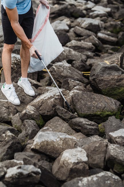 A person wearing a light blue shirt, black shorts, and white sneakers is collecting debris from a rocky outdoor area using a grabber tool while holding a large white plastic bag. The rocks are irregular in shape, varying in size, and predominantly dark gray with some lighter patches and patches of green moss or algae. The scene takes place on a natural terrain, possibly near a shoreline or riverbank, with a background of uneven rocks and natural landscape. The person appears to be engaged in clearing or collecting waste or debris, which is consistent with activities related to rubbish removal or environmental cleanup. The lighting is natural, suggesting an outdoor setting during daylight. The image conveys an act of outdoor waste collection within a rugged natural environment, consistent with private or independent waste disposal efforts.
