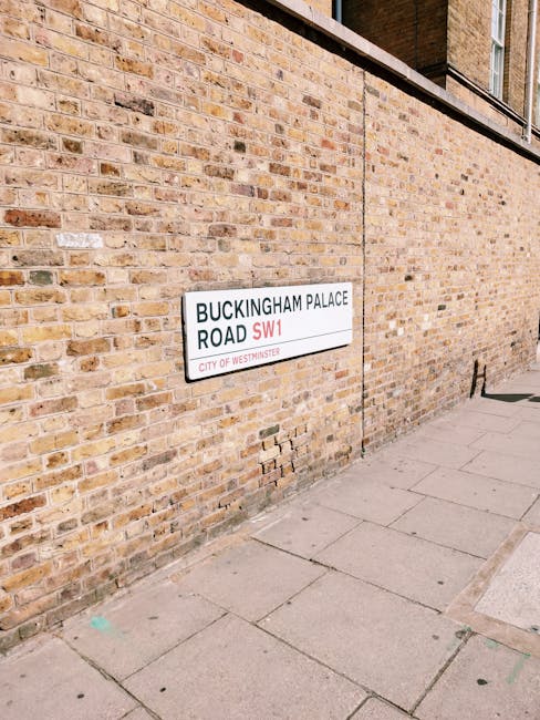 A close-up image of a white rectangular street sign mounted on a brick wall on a pavement. The sign displays the text 'BUCKINGHAM PALACE ROAD SW1' with 'SW1' in red and the remaining text in black, indicating a location in Westminster. Beneath the main address, it reads 'CITY OF WESTMINSTER' in smaller font. The brick wall has a warm, earthy tone with some visible mortar lines and slight variation in brick coloration, ranging from light beige to darker brown. The wall's surface appears weathered with some minor chips and uneven textures. The pavement in front of the sign consists of rectangular concrete slabs, with a few cracks, some faintly painted markings, and natural wear evident from foot traffic. The image is illuminated by natural daylight, casting soft shadows along the wall and paving, emphasizing the textures without harsh contrasts. This setting reflects a typical urban environment where private property signage and local street identification are visibly integrated into the city’s street scene, relevant to those seeking independent waste collection or rubbish removal services close to residential or former royal premises in Westminster.